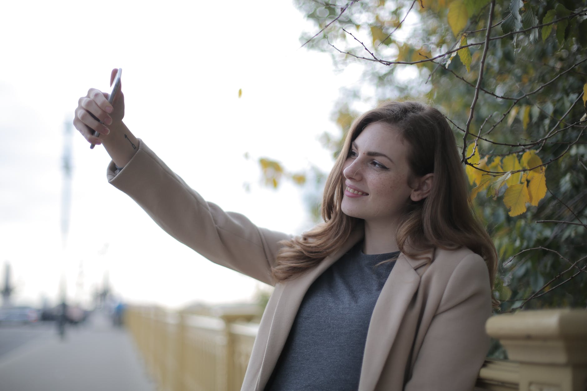 young woman in coat taking selfie with smartphone on street in overcast weather